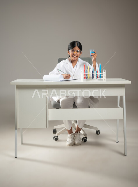 تدوين وتسجيل الملاحظات، مهن ووظائف مستقبلية، بورتريه لطفلة عربية خليجية سعودية ترتدي المعطف الطبي ونظارات الحماية تقوم بإجراء تجارب كيميائية،  العمل في مخابر التحاليل الطبية، صورة لكامل الجسم، خلفية رمادية، محتوى حساس، استخدام حساس