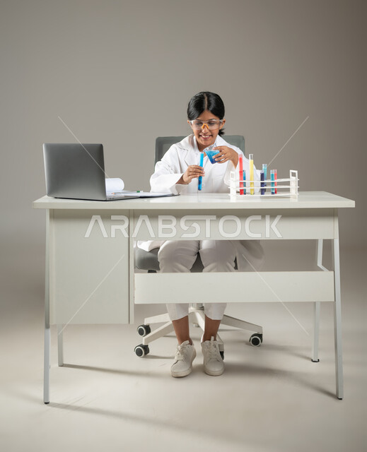 مهن ووظائف مستقبلية، دمج التكنولوجيا الحديثة في مخابر التحاليل الطبية، بورتريه لطفلة عربية خليجية سعودية ترتدي المعطف الطبي ونظارات الحماية تقوم بإجراء تجارب كيميائية باستعمال أنابيب الاختبار،  صورة لكامل الجسم، خلفية رمادية، محتوى حساس، استخدام حساس