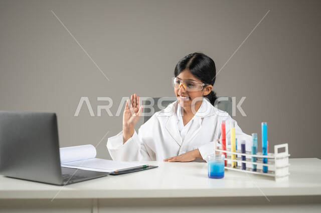 دمج التكنولوجيا في مخابر التحاليل الطبية، بورتريه مقرب لطفلة عربية خليجية سعودية ترتدي المعطف الأبيض ونظارات الحماية تقوم بالتحدث إلى شخص ما عبر الانترنت، إجراء تجارب كيميائية باستعمال أنابيب الاختبار، مهن ووظائف مستقبلية، خلفية رمادية، محتوى حساس، استخدام ساس