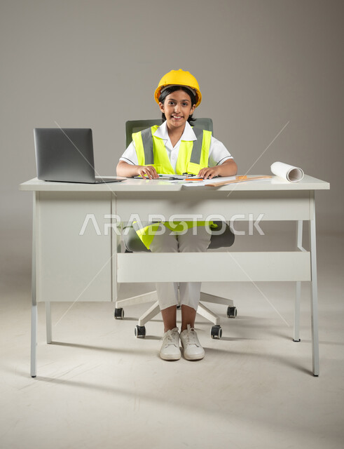 دمج التكنولوجيا الحديثة في المجال الهندسي, بورتريه لفتاة عربية خليجية سعودية ترتدي خوذة وسترة الحماية تجلس على المكتب وأمامها مخططات هندسية تنظر إلى الكاميرا بإيماءات البهجة والتفاؤل، مهن ووظائف مستقبلية، صورة لكامل الجسم، خلفية رمادية، محتوى حساس، استخدام حساس