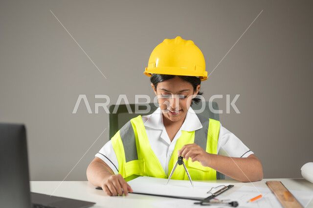 البحث عن مهنة المستقبل، ايماءات البهجة والسرور أثناء إنجاز المهام, بورتريه مقرب لطفلة عربية خليجية سعودية ترتدي خوذة وسترة الحماية تقوم باستعمال الفرجار في رسم المخططات الهندسية، دمج التقنيات الحديثة في المجال الهندسي، خلفية رمادية، محتوى حساس، استخدام حساس