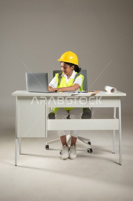 اختيار مهنة المستقبل منذ الصغر، بورتريه  لطفلة عربية خليجية سعودية ترتدي سترة وخوذة الحماية تجلس على المكتب تقوم باستعمال اللابتوب في رسم مخططات هندسية معمارية, صورة لكامل الجسم، خلفية رمادية، صورة لكامل الجسم، استخدام حساس، محتوى حساس