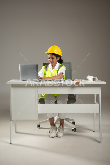 تحديد مهنة المستقبل منذ الصغر، دمج الأجهزة التقينة الحديثة في المجال الهندسي، بورتريه  لطفلة عربية خليجية سعودية ترتدي سترة وخوذة الحماية تجلس على المكتب تقوم باستعمال اللابتوب في رسم المخططات الهندسية, خلفية رمادية، صورة لكامل الجسم، استخدام حساس، محتوى حساس