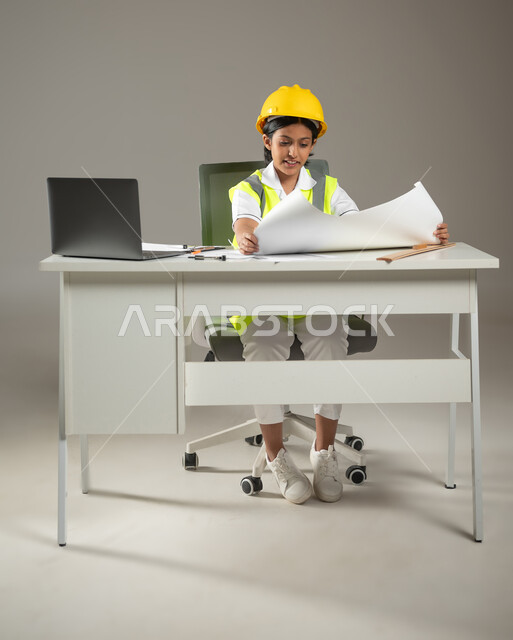طموحات الأطفال في مهنة المستقبل، إنجاز مخططات البناء، دمج التكنولوجيا الحديثة في الأعمال الهندسية، بورتريه لطفلة عربية خليجية سعودية ترتدي سترة وخوذة الحماية تقوم بمتابعة العمل خلف مكتب، صورة لكامل الجسم، خلفيه رماديه، محتوى حساس، استخدام حساس