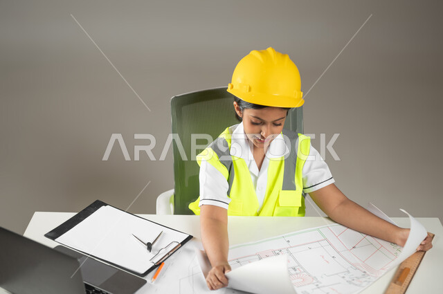 الاندماج في إنجاز مخططات البناء، مهن ووظائف مستقبلية، دمج التكنولوجيا الحديثة في الأعمال الهندسية، بورتريه مقرب لطفلة عربية خليجية سعودية ترتدي سترة وخوذة الحماية تقوم بمتابعة العمل خلف مكتب هندسي، خلفيه رماديه، محتوى حساس، استخدام حساس