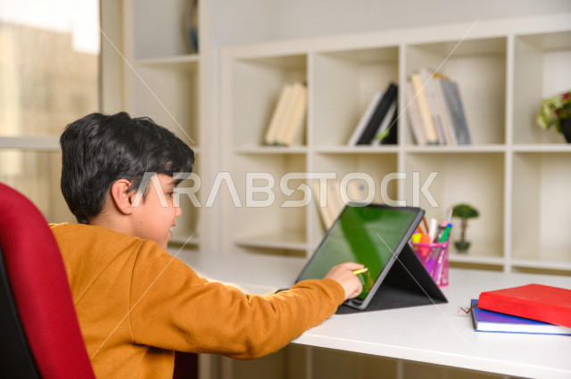 A Saudi Arabian Gulf student studies at home using the tablet with a green screen, Chroma, follows the online lessons via the laptop, distance learning, the concept of online education, studying through an educational platform for distance learning, self-