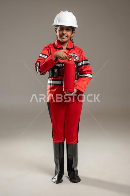 Firefighter concept for children, portrait of a Saudi Arabian Gulf girl wearing a helmet, firefighter protection jacket, and special fire-resistant shoes, standing and holding a fire extinguisher in her hand, looking at the camera with a smile, gray background, full-length body, sensitive content