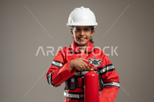 Passion for helping people and volunteering in rescue operations, portrait of a Saudi Gulf Arab girl wearing a helmet and a firefighter’s protective vest, standing and holding a fire extinguisher in her hand looking at the camera with gestures of pleasure, civil defense and civil protection, gray background, sensitive content