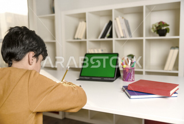 تلميذ سعودي خليجي يدرس في المنزل باستخدام جهاز التابلت بشاشة خضراء كروما ، يتابع حصص الاونلاين عن طريق الحاسوب المحمول  ، التعلم عن بعد ، مفهوم التعليم عبر الانترنت ،الدراسة من خلال منصة تعليمية للتعلم عن بعد  ، العزلة الذاتية .