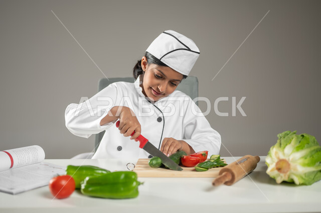 إعداد الوصفات الشهية اللذيذة وتجربتها, بورتريه لطفلة عربية خليجيه سعودية ترتدي لباس الطباخ الكامل مع القبعة البيضاء تمسك بيدها السكين وتقطع الخضار بحرص حتى لا تؤذي نفسها, تنمية مهارة الطهي والطبخ عند الأطفال,  مهن مستقبلية, خلفية رمادية, محتوى حساس