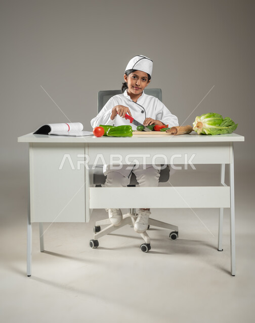 أزياء رسمية تعبر عن المهنة, بورتريه لطفلة عربية خليجيه سعودية بزي الشيف المحترف تجلس وراء طاولة الطعام وتقوم بنقطيع الخضروات الطازجة, النر إلى الكاميرا بإيماءات السعادة,  إعدداد الوصفات اللذيذة والطعام الصحي, خلفية رمادية, محتوى حساس