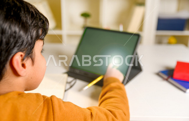 تلميذ سعودي خليجي يدرس في المنزل باستخدام جهاز التابلت بشاشة خضراء كروما ، يتابع حصص الاونلاين عن طريق الحاسوب المحمول  ، التعلم عن بعد ، مفهوم التعليم عبر الانترنت ،الدراسة من خلال منصة تعليمية للتعلم عن بعد  ، العزلة الذاتية .