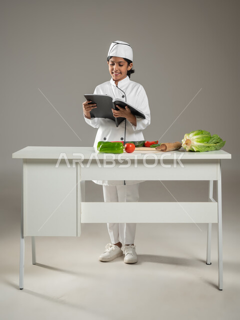 متابعة الخطوات اللازمة من كتاب وصفات الطهي الشهي, فن الطبخ وتحضير الطعام, بورتريه لطفلة عربية خليجيه سعودية ترتدي لباس الطباخ الكامل مع القبعة البيضاء تقرأ الكتاب بإيماءات السعادة, خلفية رمادية, جسم بالطول الكامل, محتوى حساس