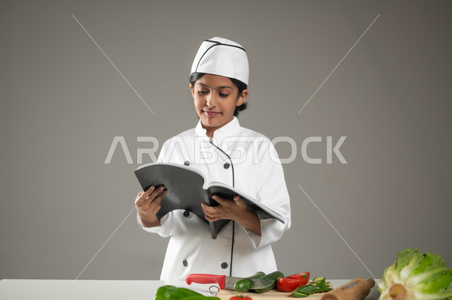 Reading a book of food recipes in order to learn innovative ways to make different foods, a portrait of a Saudi Arabian Gulf girl wearing a full chef’s uniform with a white hat, looking at the book with gestures indicating discovery, the art of cooking and cooking, healthy dishes and calories, gray background, sensitive content