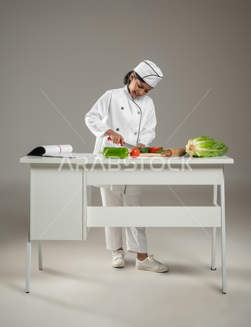 احتراف تحضير الطعام, تنمية الهوايات والمواهب منذ الصغر, بورتريه لطفلة عربية خليجيه سعودية بالزي الخاص للشيف تقوم بتقطيع الخضار بحذر لإعداد السلطة الشهية, فن الطبخ والطهي, خلفية رمادية, جسم بالطول الكامل, محتوى حساس