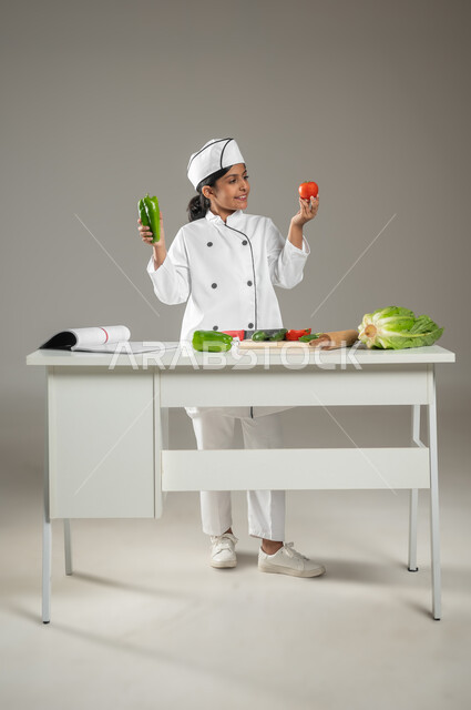 المتعة في تحضير الوصفات الشهية, بورتريه لطفلة عربية خليجيه سعودية ترتدي زي الشيف مع قبعته البيضاء, تمسك بيديها الخضروات المختلفة الألوان وتقارن بينها بإيماءات الاكتشاف والسعادة, خلفية رمادية, جسم بالطول الكامل، محتوى حساس