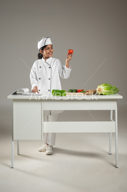 تحضير العدد اللازم من حبات الطماطم من أجل إعداد السلطة, بورتريه لطفلة عربية خليجيه سعودية ترتدي لباس الطباخ الكامل مع القبعة البيضاء, وجود خضروات متنوعة  وكتاب الطهي على الطاولة, تقديم أشهى الأطباق, خلفية رمادية, جسم بالطول الكامل، محتوى حساس