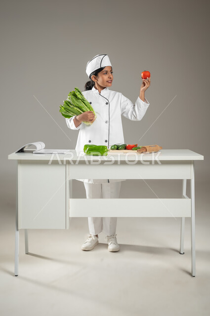 الاستعداد والبدء بتجهيز الوصفة, التأكد من سلامة جميع الخضروات, بورتريه لطفلة عربية خليجيه سعودية ترتدي لباس الطباخ الكامل مع القبعة البيضاء وأمامها على الطاولة جميع المكونات, تقطيع الخصار وإعداد السلطة, خلفية رمادية, جسم بالطول الكامل، محتوى حساس