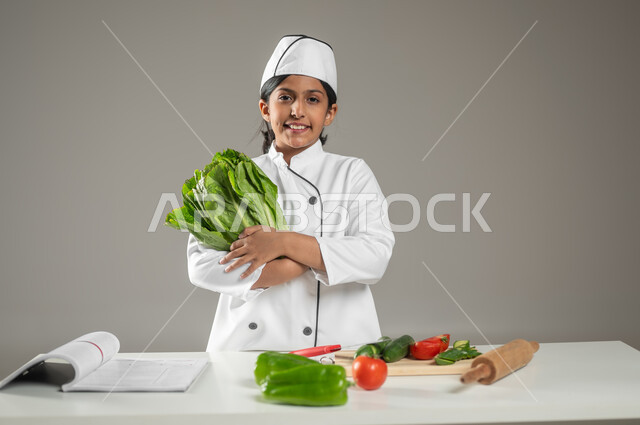 النظر إلى الكاميرا بإيماءات السرور والإمساك بمكونات الوصفة, بورتريه لطفلة عربية خليجيه سعودية ترتدي لباس الطباخ مع القبعة البيضاء تقف وبيدها الخس, الخضروات الشهية المتنوعة الألوان والأشكال موضوعة على الطاولة المطبخية, إعداد وصفات الطعام وتحضيرها, خلفية رمادية, محتوى حساس