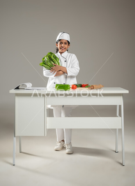 الوقوف خلف طاولة المطبخ في إشارة إلى الاستعداد والبدء بتجهيز الوصفة, بورتريه لطفلة عربية خليجيه سعودية ترتدي لباس الطباخ الكامل مع القبعة البيضاء وأمامها على الطاولة خضروات متنوعة, تقطيع الخصار وإعداد السلطة, خلفية رمادية, جسم بالطول الكامل، محتوى حساس