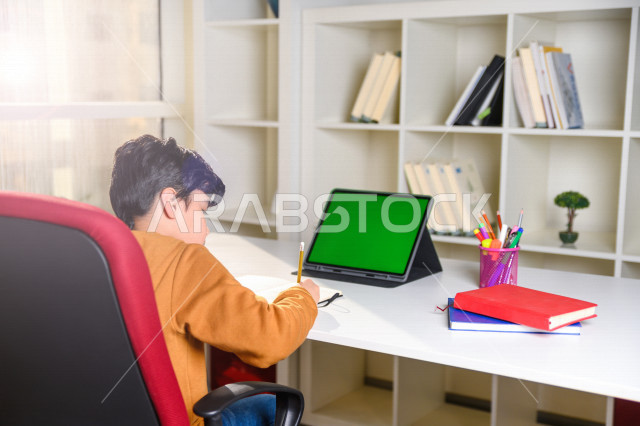 تلميذ سعودي خليجي يدرس في المنزل باستخدام جهاز التابلت بشاشة خضراء كروما ، يتابع حصص الاونلاين عن طريق الحاسوب المحمول  ، التعلم عن بعد ، مفهوم التعليم عبر الانترنت ،الدراسة من خلال منصة تعليمية للتعلم عن بعد  ، العزلة الذاتية .
