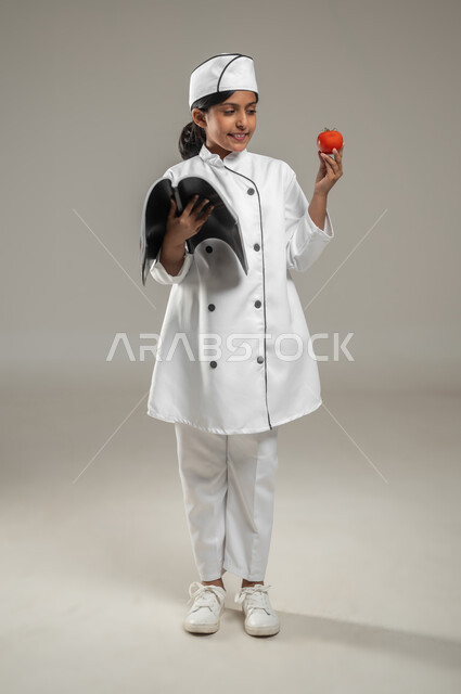 ارتداء زي الشيف والدخول إلى عالم الطهي, بورتريه لطفلة عربية خليجيه سعودية ترتدي القبعة الطباخ البيضاء مع زيه الخاص تمسك بيدها كتاب الوصفات, حمل حبة الطماطم والنظر إليها, تعليم بطرائق مبتكرة, جسم بالطول الكامل, خلفية رمادية, محتوى حساس