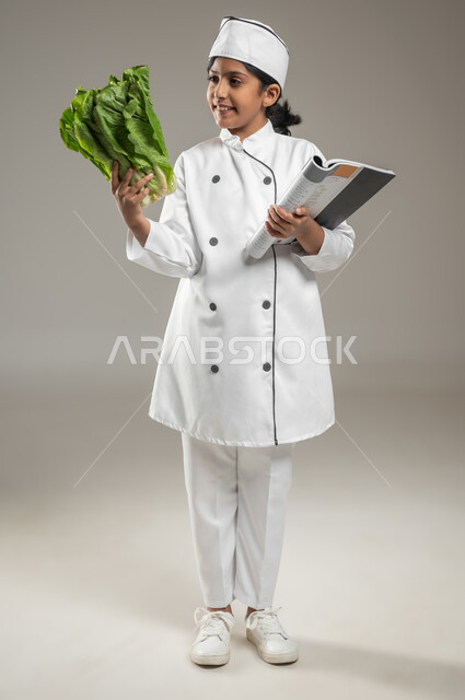 الاستمتاع بتحضير الوجبات الخفيفة كالسلطات, بورتريه لطفلة عربية خليجيه سعودية ترتدي لباس الطباخ الكامل مع القبعة البيضاء تمسك بيد الخس وباليد الأخرى تحمل كتاب الخطوات لإعداد الوصفة, النظر إلى أوراق الخس بتمعن, فن الطبخ والطهي, خلفية رمادية, محتوى حساس