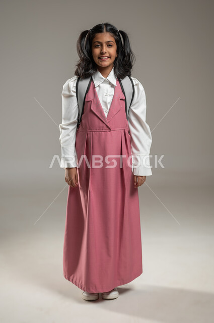 النظر إلى الكاميرا بإيماءات السرور, الوقوف بشكل مستقيم يدل على الثقة بالنفس, بورتريه لتلميذة عربية خليجية سعودية ترتدي زيها الرسمي وحقبيتها المدرسية, دروس و واجبات منزلية, جسم بالطول الكامل, خلفية رمادية, محتوى حساس