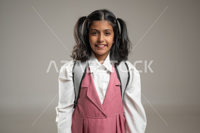 The role of education in the renaissance of the Kingdom of Saudi Arabia, new and innovative teaching methods, effective learning in Saudi society, a portrait of a Saudi Gulf Arab student wearing her school uniform and looking at the camera with happy gestures, lessons and homework, gray background, sensitive content