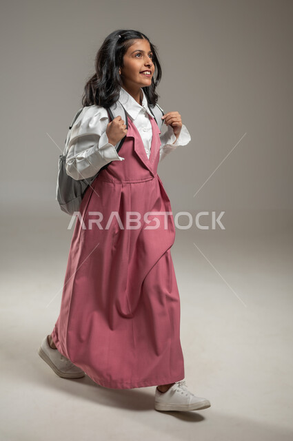 النظر إلى الأفق والتأمل بتحقيق أحلام النجاح,السعي نحو طلب العلم, بورتريه بخلفية رمادية لتلميذة عربية خليجية سعودية تحمل حقيبتها المرسية  وترتدي الزي الرسمي المخصص, وظائف وواجبات منزلية, جسم بالطول الكامل, محتوى حساس