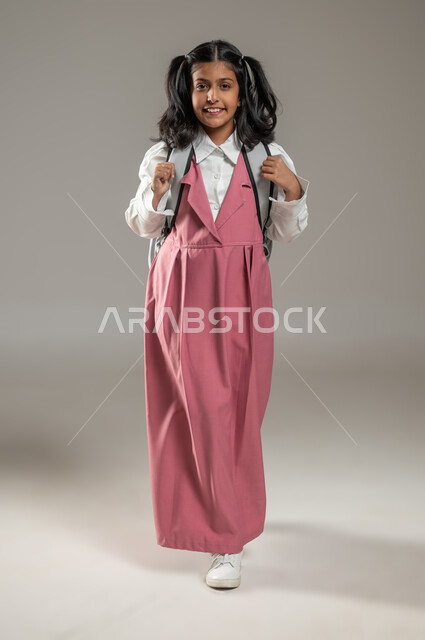 السعي نحو النجاح والتفوق, الجد والاجتهاد والدراسة بشكل جيد, التعلم الفعال والمشاركة مع الطلاب, بورتريه بخلفية رمادية لتلميذة عربية خليجية سعودية تقف وتنظر إلى الكاميرا بإيماءات السرور, دروس و واجبات منزلية, جسم بالطول الكامل, محتوى حساس