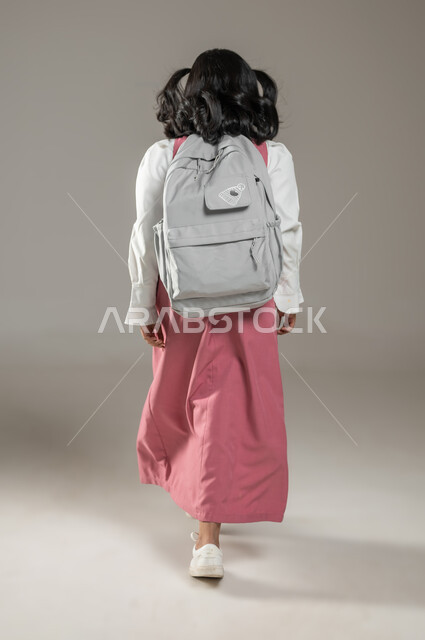 المدارس والتدريس في المملكة العربية السعودية, طرائق تدريس جديدة ومبتكرة, بورتريه من الخلف لتلميذة عربية خليجية سعودية ترتدي زيها المدرسي وتحمل حقبيتها فوق ظهرها وتتجه نحو المدرسة,الجد والاجتهاد والسعي نحو النجاح, خلفية رمادية, جسم بالطول الكامل, محتوى حساس