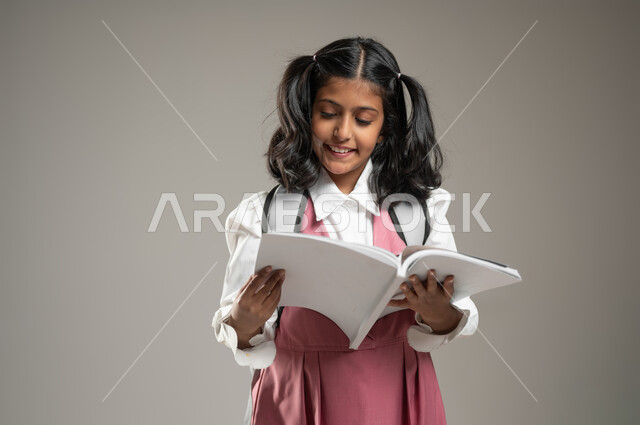 هواية القراءة والمطالعة, مراجعة مواضيع الكتب المدرسية,طرائق تدريس حديثة, التعلم الفعال والمشاركة مع الطلاب, بورتريه لتلميذة عربية خليجة سعودية ترتدي الزي الرسمي المخصص للمدرسة تمسك بيدها كتابها وتقرأ منه, دراسة وواجبات منزلية, خلفية رمادية, محتوى حساس