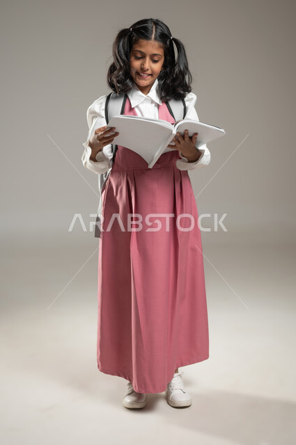 الجد والاجتهاد في التعلم, عام دراسي جديد, بورتريه لتلميذة عربية خليجة سعودية ترتدي الزي الرسمي المخصص للمدرسة تحمل بيدها كتاب تقوم بقرائته, دراسة وواجبات منزلية, قراءة ومطالعة وتفوق, خلفية رمادية, جسم بالطول الكامل, محتوى حساس