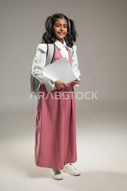 الوقوف باستعداد وجاهزية لاستقبال العام الدراسي, التعلم الفعال والمشاركة مع الطلاب, بورتريه لتلميذة عربية خليجة سعودية ترتدي الزي الرسمي المخصص تحمل بيدها مجموعة من الأوراق, دراسة وواجبات منزلية, طرائق تدريس حديثة, جسم بالطول الكامل, خلفية رمادية, محتوى حساس