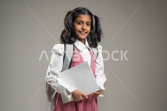 النظر إلى الكاميرا بعيون مليئة بالتفاؤل والثقة, طرائق تدريس حديثة, التعلم الفعال والمشاركة مع الطلاب, بورتريه لتلميذة عربية خليجة سعودية ترتدي الزي الرسمي المخصص للمدرسة تمسك بيدها أوراق, دراسة وواجبات منزلية, خلفية رمادية, محتوى حساس