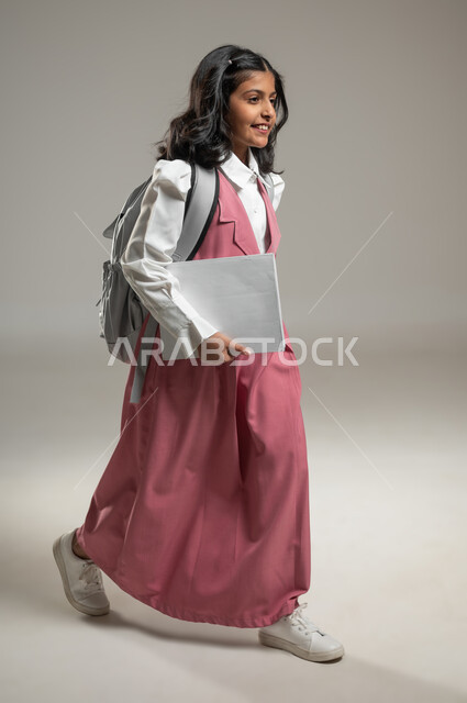تطور التعليم في المملكة العربية السعودية, التعلم الفعال والمشاركة مع الطلاب, بورتريه لتلميذة عربية خليجة سعودية ترتدي الزي الرسمي المخصص للمدرسة وتمشي بحطوات واثقة نحو بناء مستقبلها, دراسة وواجبات منزلية, خلفية رمادية, جسم بالطول الكامل, محتوى حساس