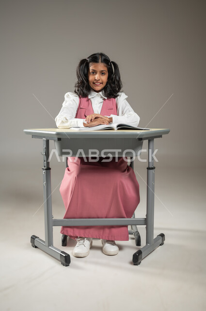 Discipline and adherence to school rules, Portrait of a Saudi Gulf Arab student sitting in her assigned seat, wearing the official school uniform and opening her books in front of her, lessons and homework, new and innovative teaching methods, effective learning and participation with students, gray background, sensitive content