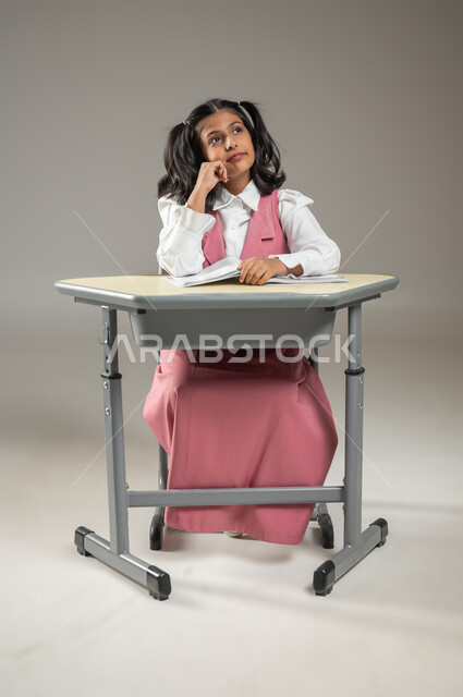 Following dreams with all determination and resolve, looking at the horizon with eyes hoping to achieve success and excellence, a portrait of a Saudi Gulf Arab student sitting in her seat and wearing the official school uniform, the development of education in Saudi Arabia, gray background, sensitive content