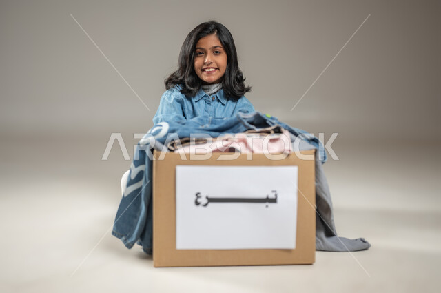 Looking at the camera with eyes full of gratitude and joy with a big smile, the joy of a Saudi Gulf Arab girl getting some clothes, giving Eid clothes to the needy, charities and humanitarian initiatives, portrait with a gray background, charitable volunteer initiatives, sensitive content