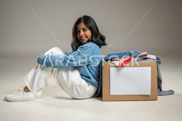 إهداء ملابس العيد للمحتاجين, الجلوس بجانب صندوق التبرعات تعبيراً عن الفرح، جمعيات خيرية ومبادرات انسانية، بورتريه بخلفية رمادية لطفلة عربية خليجية سعودية ترتدي زي كاجوال لطيف, مبادرات تطوعية خيرية, محتوى حساس