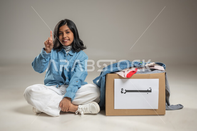 Sitting next to the donation box and raising the hand indicating the joy that the child is experiencing, a portrait with a gray background of a Saudi Arabian Gulf girl wearing a casual outfit, looking at the camera and smiling, Islamic associations in Saudi Arabia, sensitive content
