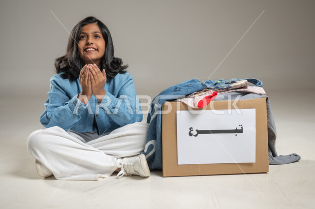 الحصول على صندوق مليء بالملابس من التبرعات الخيرية، بورتريه بخلفية رمادية لطفلة عربية خليجية سعودية ترتدي زي كاجوال تقوم برفع كفيها والدعاء لأهل الخير الذين قدموا لها الصندوق, جمعيات خيرية ومبادرات انسانية, محتوى حساس