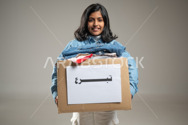 Charitable organizations and humanitarian initiatives, collecting and distributing donations to the poor and needy, a portrait with a gray background of a Saudi Gulf Arab girl wearing a nice casual outfit carrying a box of clothes in order to distribute them to the needy, with gestures of joy, sensitive content
