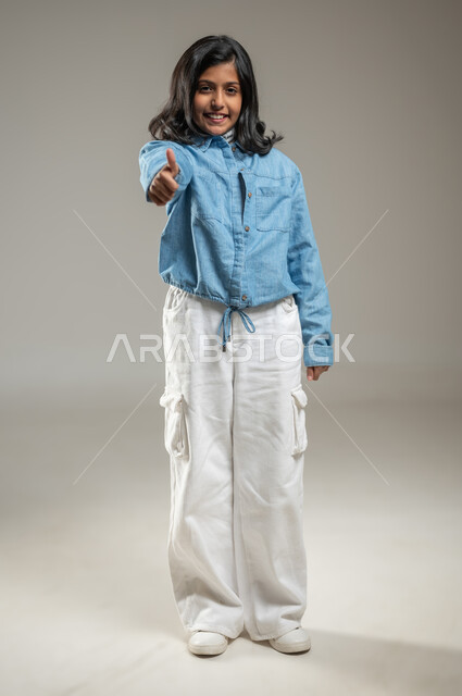 رفع اليد بإشارة تدل على السعادة,الوقوف والنظر بشكل مباشر إلى الكاميرا, إيماءات الوجه تدل على الفرح والسرور, بورتريه لبنت لطيفة عربية خليجة سعودية ترتدي زي كاجوال جميل, جسم بالطول الكامل, خلفية رمادية, محتوى حساس