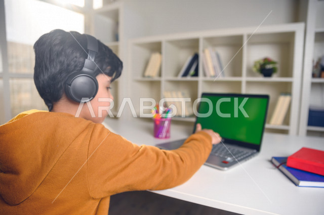 تلميذ سعودي خليجي يدرس في المنزل باستخدام جهاز التابلت بشاشة خضراء كروما ، يتابع حصص الاونلاين عن طريق الحاسوب المحمول  ، التعلم عن بعد ، مفهوم التعليم عبر الانترنت ،الدراسة من خلال منصة تعليمية للتعلم عن بعد  ، العزلة الذاتية .