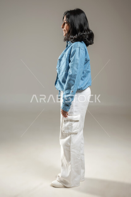 الوقوف بشكل مستقيم والنظر إلى الأمام، بورتريه  من الجانب لفتاة عربية خليجية سعودية أنيقة ترتدي قميص جميل أزرق اللون وبنطال أبيض, الشعر الأسود المنساب على كتفيها يظهر لامعاً من انعكاس الضوء عليه، جسم بالطول الكامل, خلفية رمادية, محتوى حساس, استخدام حساس