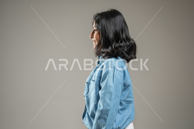 التزيين والتجمل والإهتمام بالمظهر الخارجي، بورتريه  من الجانب لفتاة عربية خليجية سعودية أنيقة ترتدي قميص جميل أزرق اللون وبنطال أبيض, الشعر الأسود المنساب على كتفيها يظهر لامعاً من انعكاس الضوء عليه، خلفية رمادية, محتوى حساس, استخدام حساس