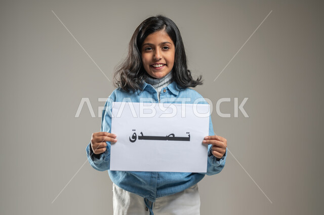 Carrying out voluntary charitable initiatives, launching campaigns that explain the importance of charity in the life of a Muslim person, a portrait of a Saudi Gulf Arab girl holding a white board with the words charity written on it, gestures of joy and happiness, gray background, sensitive content, sensitive use