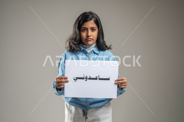 The need for help and to provide protection and care, a portrait of a Saudi Arabian Gulf girl holding in her hands a paper written in bold, Help me, looking at the camera with an expression of sadness, raising awareness about the dangers of child abuse, gray background, sensitive content, sensitive use